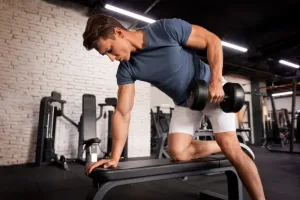 Hombre realizando remo con mancuerna en banco plano en gimnasio, ejercicio ideal para fortalecer la espalda y mejorar la definición muscular.